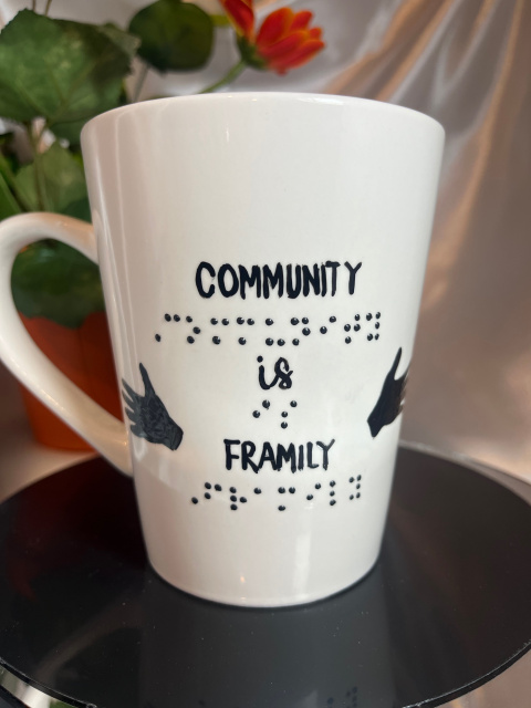 Shown- Front of 14 ounce white mug with message in raised braille and script with two outreaching hands in black