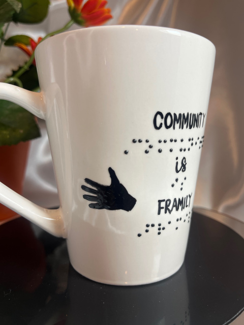 Shown- Left side of 14 ounce white mug with message in raised braille and script with outreaching hand in black
