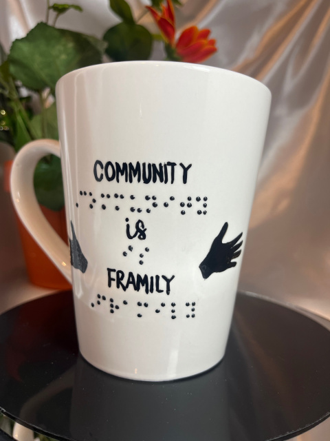 Shown- Right side of 14 ounce white mug with message in raised braille and script with outreaching hand in black